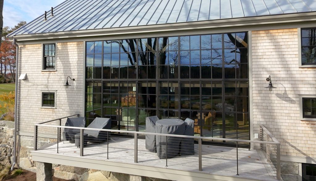 A wide-angle exterior shot reveals a cedar-shingled building with a standing-seam metal roof and a massive black-mullioned glass wall featuring double French doors with brass handles, which reflects a dense forest and a bright sky while overlooking a raised wooden deck furnished with grey-covered patio chairs and a table set, all bordered by a modern cable railing and set against a backdrop of a lush green lawn, autumn foliage, and a distant lake.
