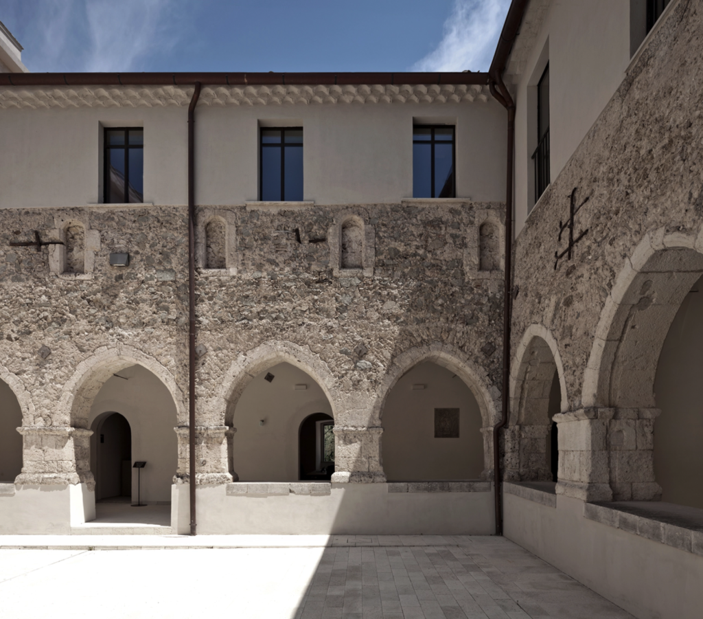 The historic "Corte storica" courtyard of the Procura della Repubblica di Catanzaro, formerly a 15th-century Franciscan military hospital, features a weathered, multi-textured stone facade punctuated by a series of pointed Gothic arches that form a colonnaded walkway, while modern slate-blue OS2 metal-framed windows are symmetrically aligned on the upper white-plastered level, all under a clear blue sky.