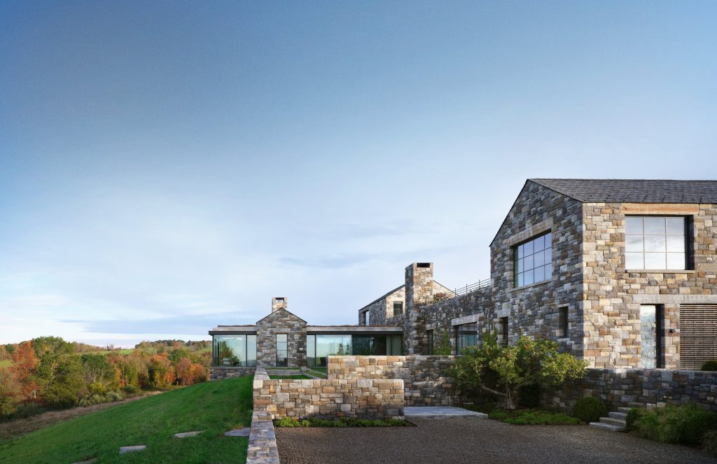 A sprawling, contemporary estate constructed from multi-toned, rustic fieldstone sits integrated into a rolling green hillside, featuring expansive curtain wall systems, clean geometric gables, and stone terrace walls that overlook a dense autumnal forest under a vast, pale blue sky