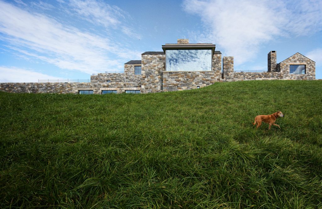A sprawling, multi-level modern residence constructed from rugged, earth-toned fieldstone sits atop a lush, verdant grassy hillside under a bright blue sky with wispy clouds, while a medium-sized brown dog trots across the foreground slope toward the right, highlighting the architectural blend of natural textures and expansive glass windows that reflect the sky