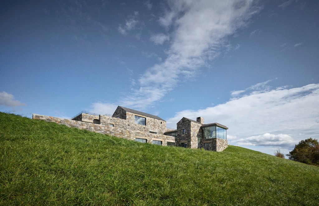 A sprawling, multi-level modern residence constructed from rugged, earth-toned fieldstone sits atop a lush, verdant grassy hillside under a bright blue sky with wispy clouds