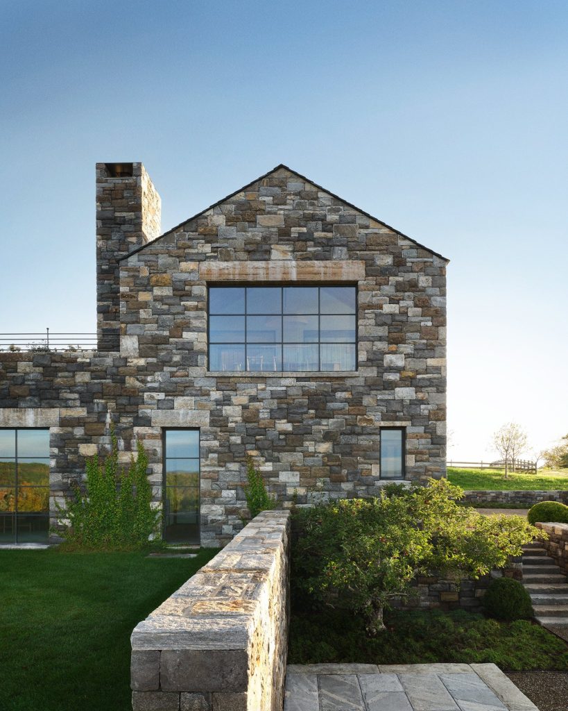 A striking, contemporary gabled structure built from textured, multi-tonal grey and tan fieldstone rises from a manicured green lawn, featuring a large, black-framed industrial window that reflects a distant horizon, a tall rectangular chimney, and a low stone retaining wall that guides the eye toward a stone staircase and lush garden greenery under a clear, bright sky.