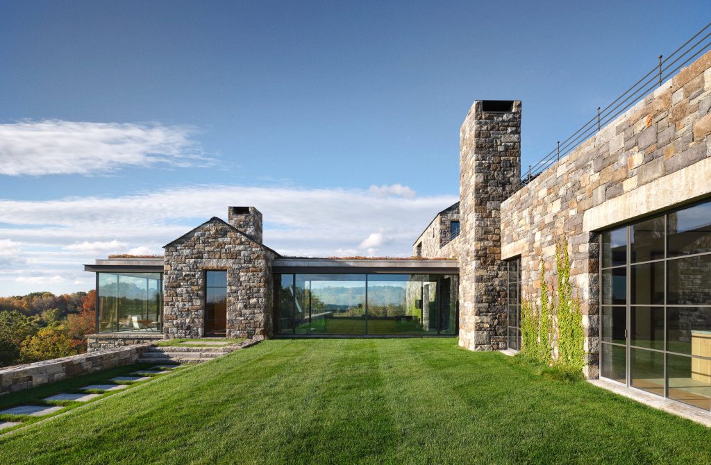 Expansive modern stone residence set on a gently sloping landscaped lawn, composed of multiple gabled volumes clad in variegated natural stone with tall rectangular chimneys, large curtain wall systems and sliding doors framed in dark steel creating strong indoor–outdoor connections, a low horizontal connector volume with a green roof element linking structures, stepped stone pathways and retaining walls integrated into the terrain, reflections of distant rolling hills and autumn foliage visible through the glazing, and a clear blue sky casting crisp daylight across the façade, emphasizing a balance of rustic materiality, contemporary geometry, and panoramic landscape integration