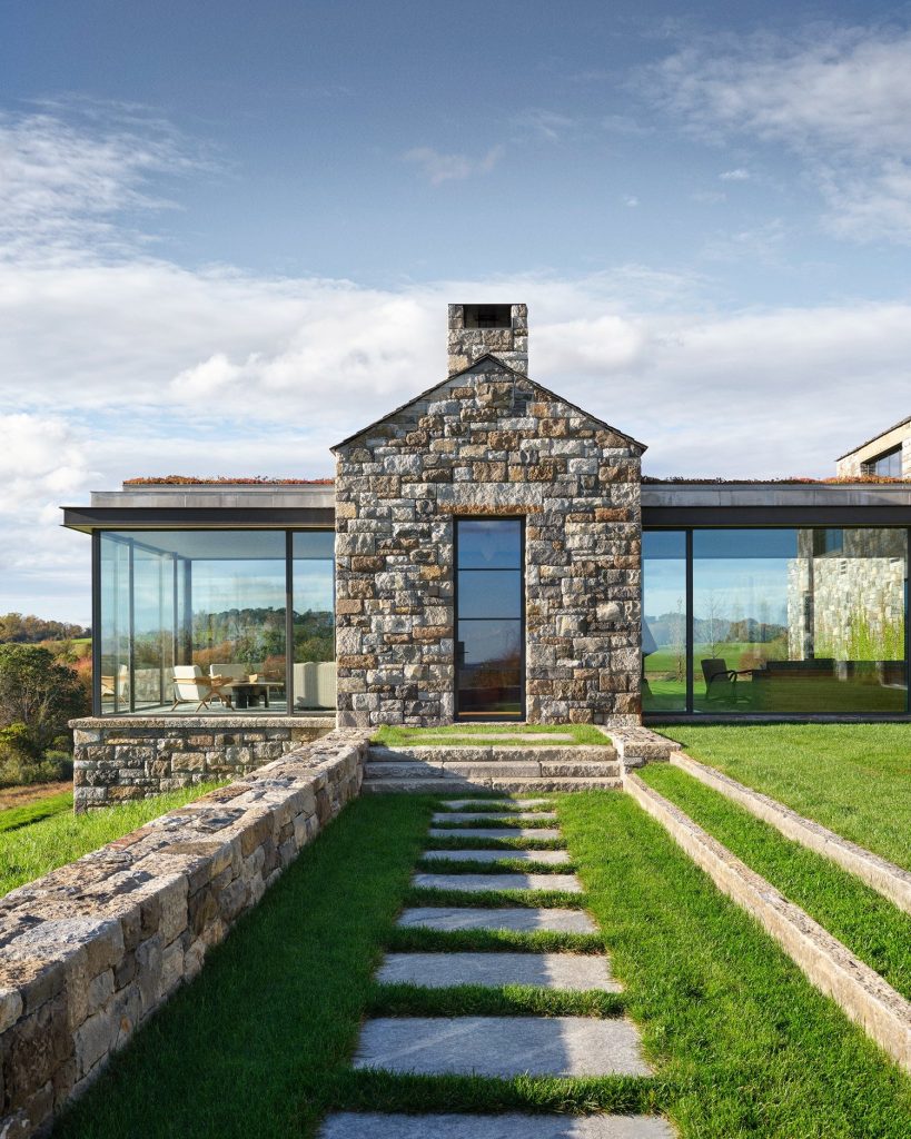 A striking, contemporary gabled structure built from textured, multi-tonal grey and tan fieldstone rises from a manicured green lawn, featuring a large, black-framed steel window that reflects a distant horizon, a tall rectangular chimney, and a low stone retaining wall that guides the eye toward a stone staircase and lush garden greenery under a clear, bright sky