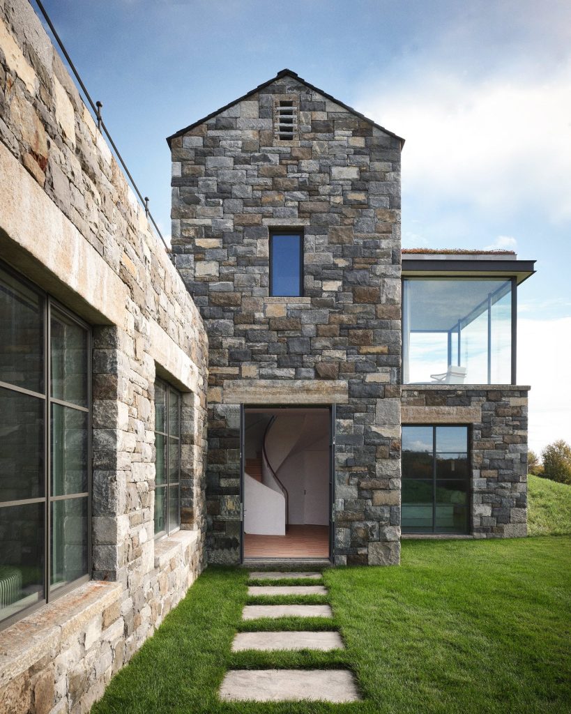 contemporary stone-clad residence featuring a central gabled volume with irregularly patterned masonry in varied gray and beige tones, tall narrow window punctuating the upper facade above a recessed glass entry door revealing a minimalist interior staircase, adjacent flat-roofed wing with expansive curtain wall system and a transparent corner revealing modern furnishings, linear stepping-stone pathway set into a neatly trimmed green lawn leading directly to the entrance, thick stone walls with deep-set windows and dark metal frames emphasizing depth and texture, subtle rooftop greenery blending the structure into the surrounding landscape, clean architectural lines merging rustic materiality with modern transparency, soft daylight illuminating the façade and highlighting the contrast between solid stone masses and reflective glass surfaces, tranquil residential setting with open sky and distant vegetation reinforcing a serene, high-end architectural aesthetic