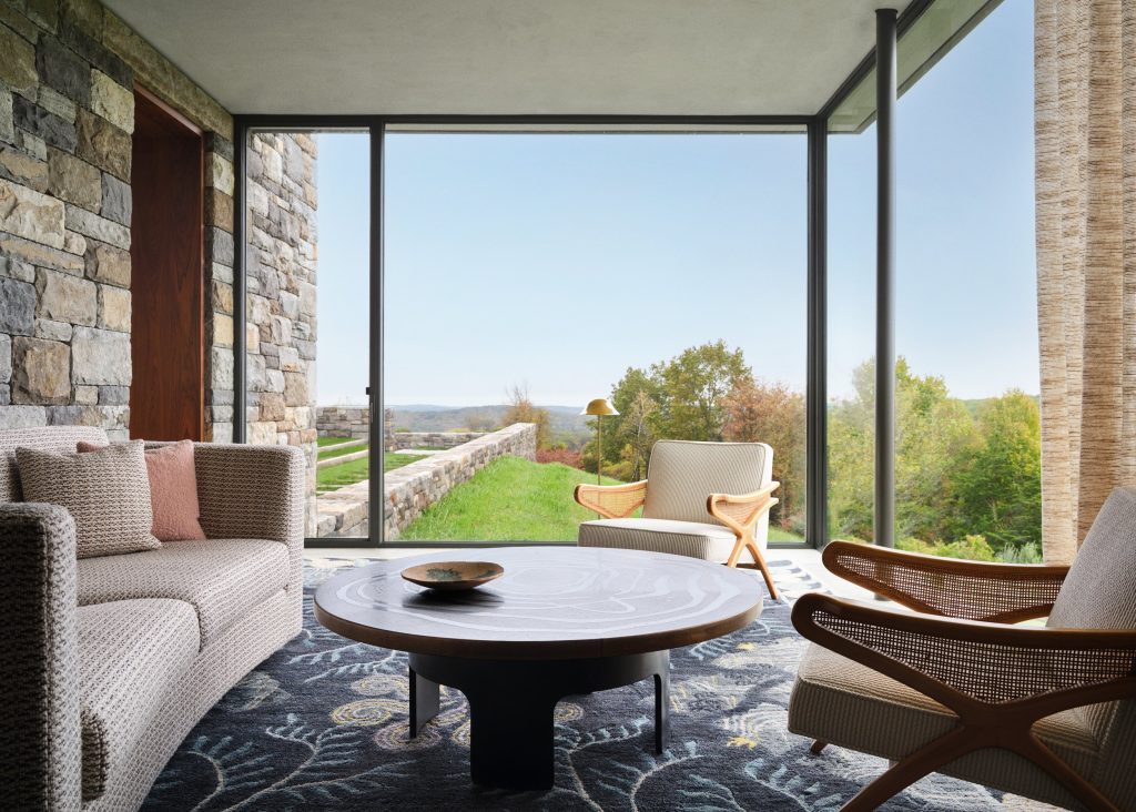 An elegant, mid-century modern sunroom features a rugged, multi-tonal bluestone accent wall, floor-to-ceiling glass windows that provide a panoramic view of a rolling green hillside and distant autumnal forest, a low-profile grey textured sofa adorned with a dusty rose pillow, two sculptural armchairs with woven cane sides and light-toned upholstery, a large circular dark wood coffee table with a swirling grain pattern centered on a plush navy blue area rug with intricate floral and leaf motifs, and a warm wooden doorway that opens into an adjacent space under a clean, neutral-toned ceiling.