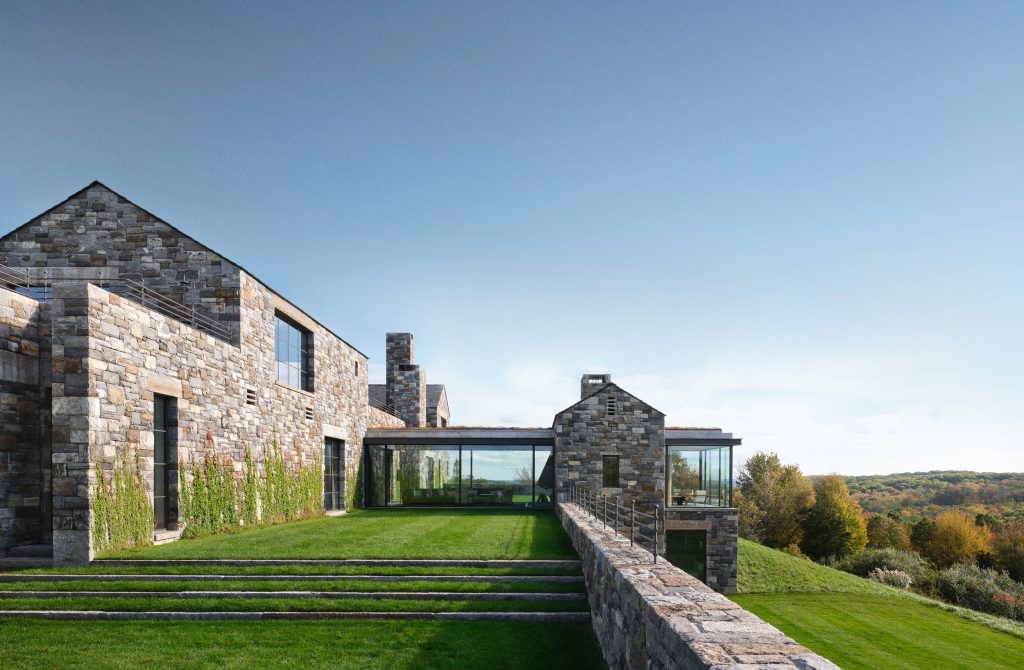 A sprawling, multi-level modern residence constructed from rugged, earth-toned fieldstone sits integrated into a lush, verdant grassy hillside under a clear blue sky, featuring a series of interconnected gabled volumes and a central glass-walled pavilion that creates a transparent bridge between the solid masonry structures, all overlooking a vast, rolling autumnal forest in the distance.