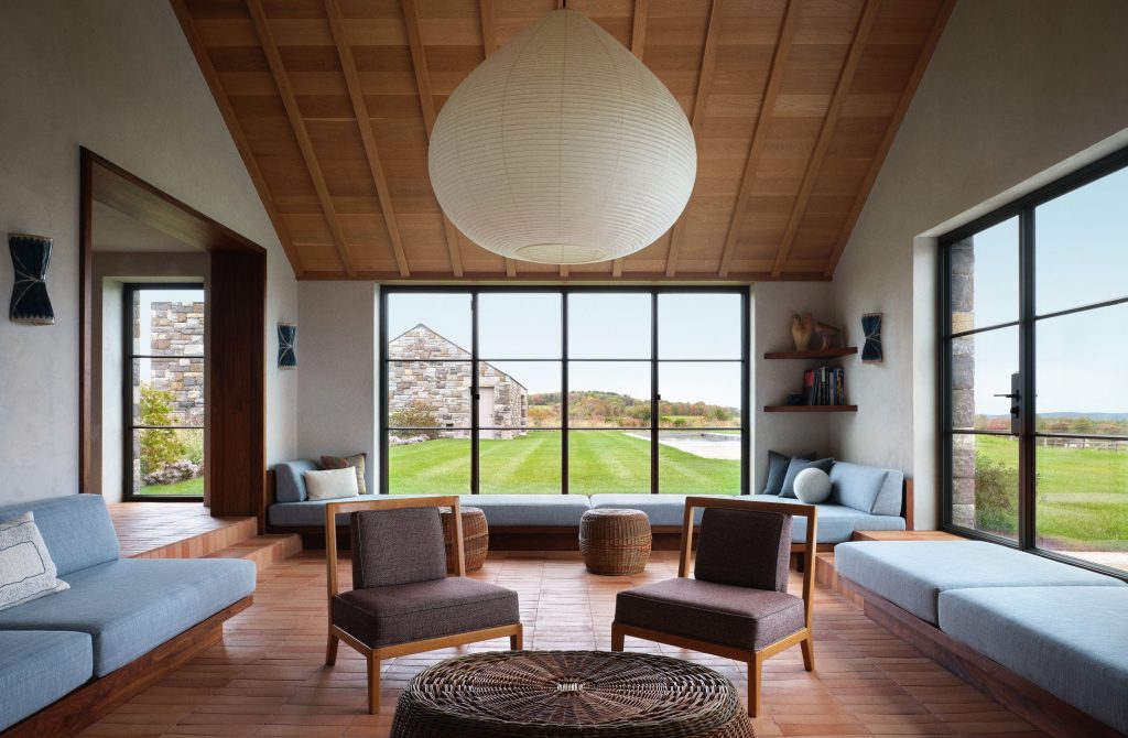 An expansive, contemporary living room features a warm, wood-slatted gabled ceiling with a massive, spherical paper lantern suspended centrally, light blue built-in daybeds with plush cushions that line the perimeter of the terracotta-tiled floor, two low-profile wooden armchairs with dark textured upholstery facing a large circular woven coffee table, and massive black-framed industrial windows that frame a manicured green lawn and a distant stone gabled building under a soft, overcast sky.