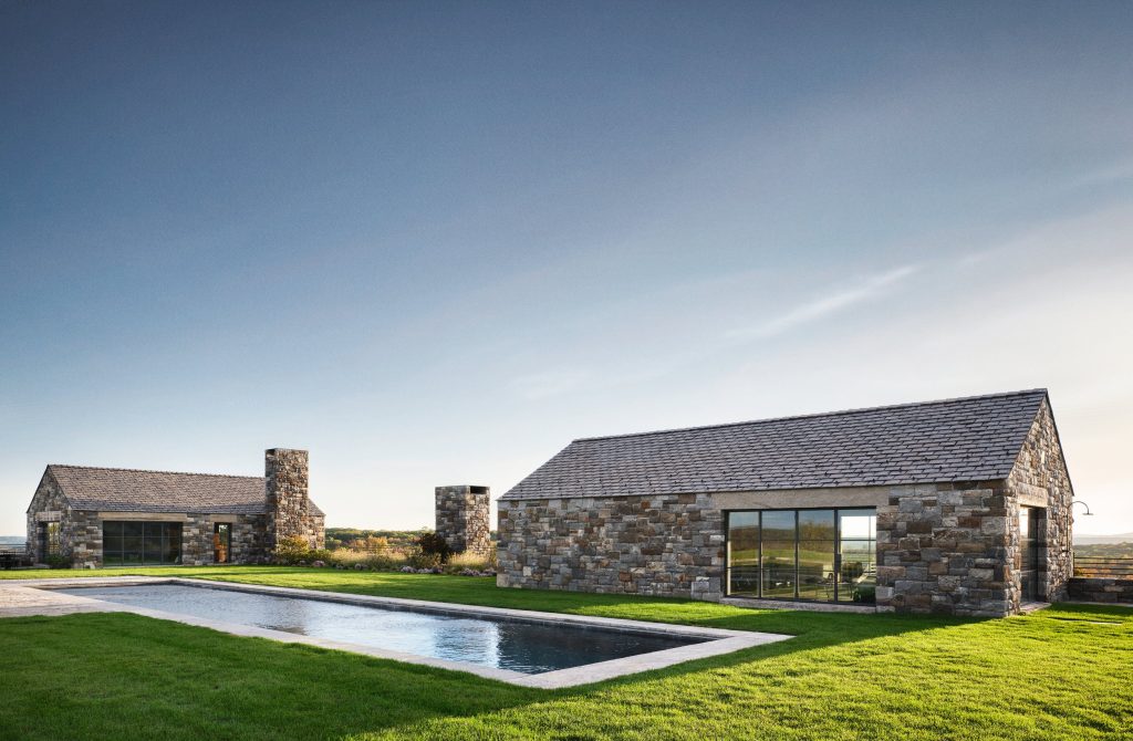 Two minimalist, gabled pavilions constructed from rustic, multi-tonal grey and tan fieldstone stand elegantly on a manicured green lawn, featuring large black-framed glass sliding doors that reflect the surrounding landscape, a rectangular stone chimney rising between the structures, and a long, rectangular lap pool with a stone border that stretches across the foreground under a vast, soft blue sky.