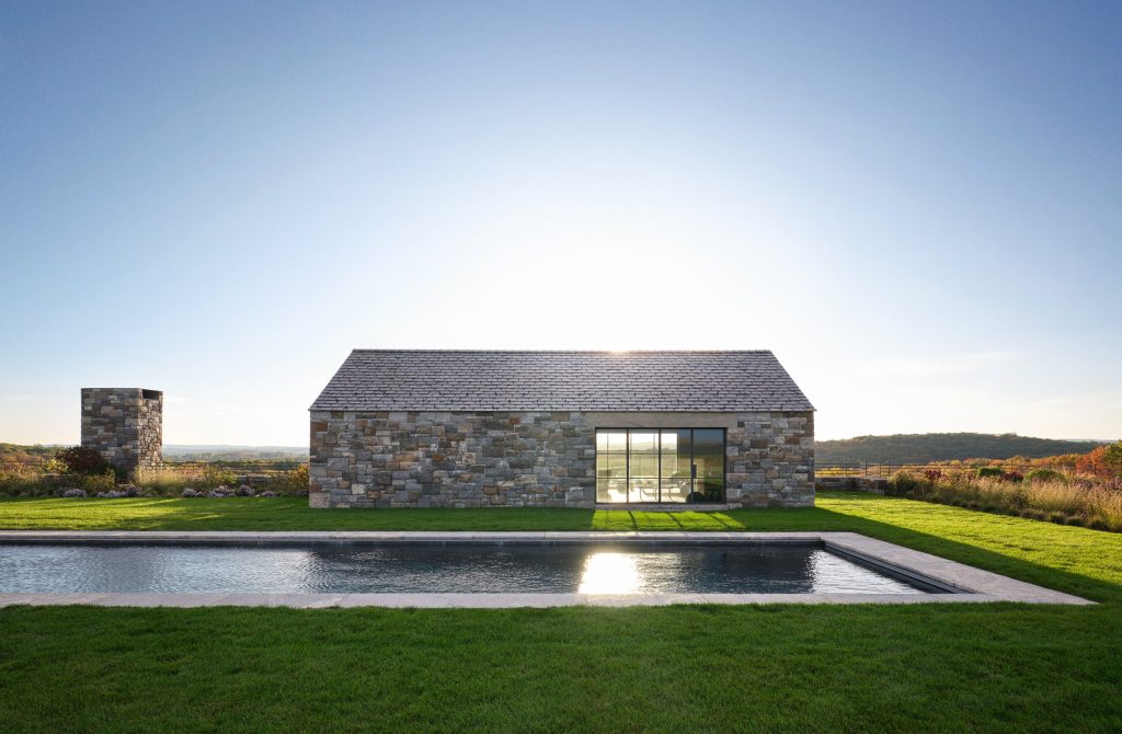 A minimalist, gabled pavilion constructed from rustic, multi-tonal grey and tan fieldstone stands elegantly on a manicured green lawn, featuring a large black-framed glass sliding door that reflects the surrounding landscape, a rectangular stone chimney rising to the left, and a long, rectangular lap pool with a stone border that stretches across the foreground under a vast, bright blue sky with the sun centered directly above the roofline