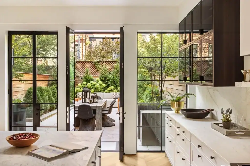 Landmark Systems NYC luxury kitchen, white marble island, open black-framed French doors, private garden terrace, outdoor dining table, wicker chairs, hydrangea landscaping, dark wood upper cabinets, marble backsplash, white lower cabinetry, brass planter, dark bowl, wooden fruit bowl, herringbone hardwood floors, NYC townhouse indoor-outdoor living.