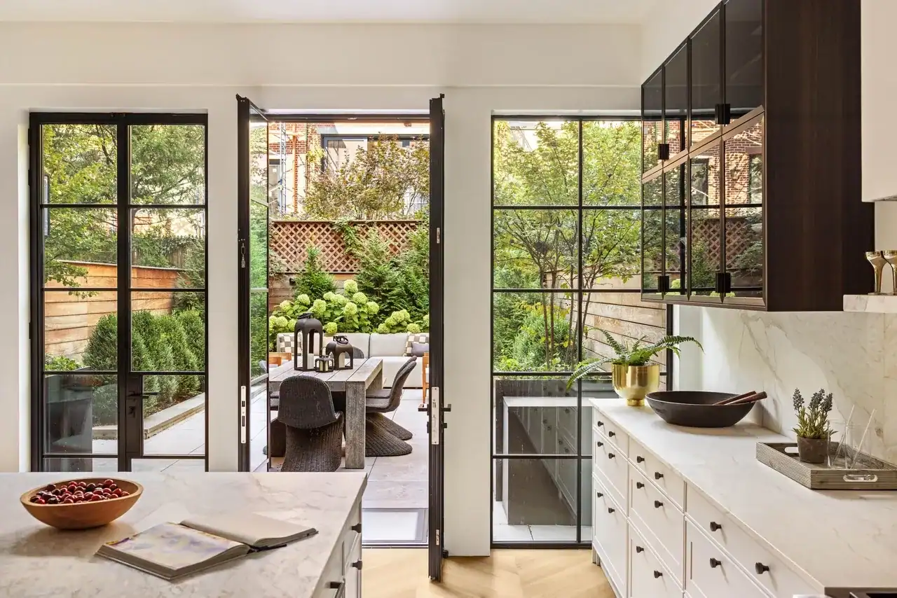 Landmark Systems NYC luxury kitchen, white marble island, open black-framed French doors, private garden terrace, outdoor dining table, wicker chairs, hydrangea landscaping, dark wood upper cabinets, marble backsplash, white lower cabinetry, brass planter, dark bowl, wooden fruit bowl, herringbone hardwood floors, NYC townhouse indoor-outdoor living.