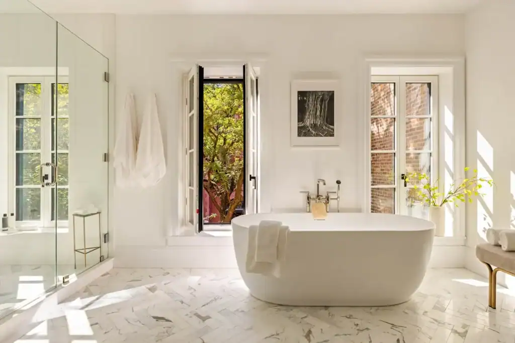 A luminous, spa-like sanctuary, defined by its pristine white aesthetic and herringbone-patterned marble flooring, centers upon a sculptural, freestanding oval bathtub positioned between two elegant white-framed windows that invite the outside in—one offering a glimpse of a verdant tree and the other revealing a classic red-brick facade—while a glass-enclosed walk-in shower with gleaming marble tiles, a minimalist black-and-white framed photograph, plush white towels, and a delicate sprig of yellow blossoms in a simple vase collectively cultivate an atmosphere of tranquil, high-end refinement.