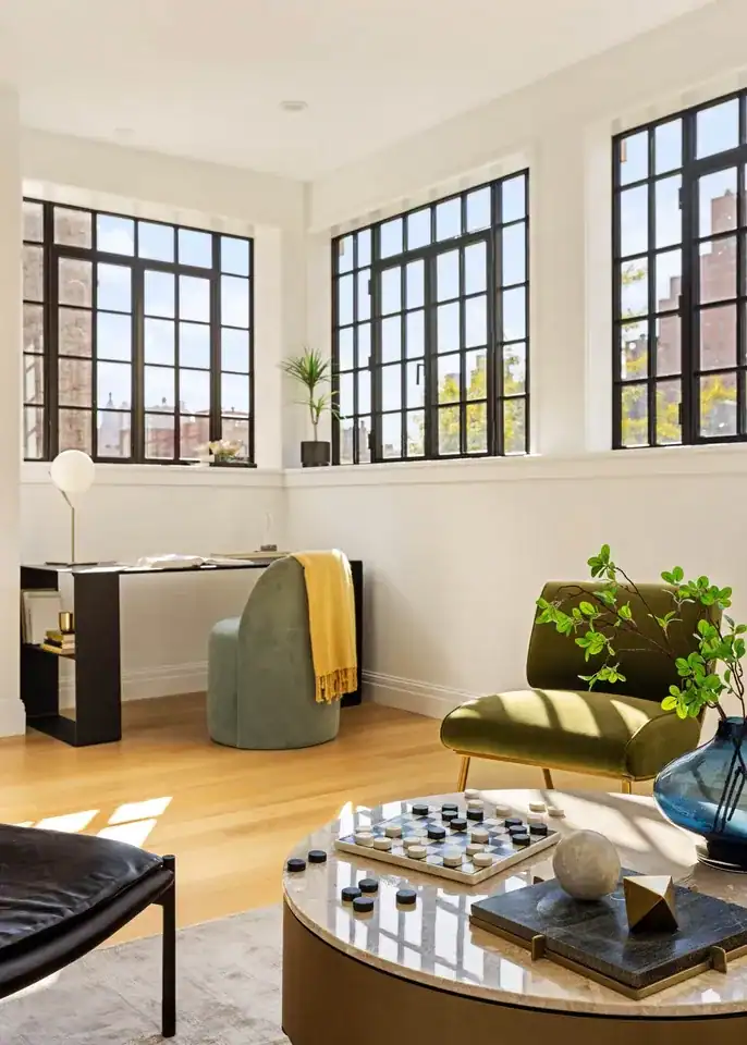 Landmark Systems NYC contemporary living room, black-framed grid windows, golden afternoon light, cream bouclé sofa, neutral toss pillows, spherical white cushion, moss-green velvet lounge chair, brass legs, silver area rug, light wood floors, gold drum coffee table, marble chessboard, blue glass vase, gold cylindrical side tables, teal magenta abstract painting, white walls, luxury interior design.