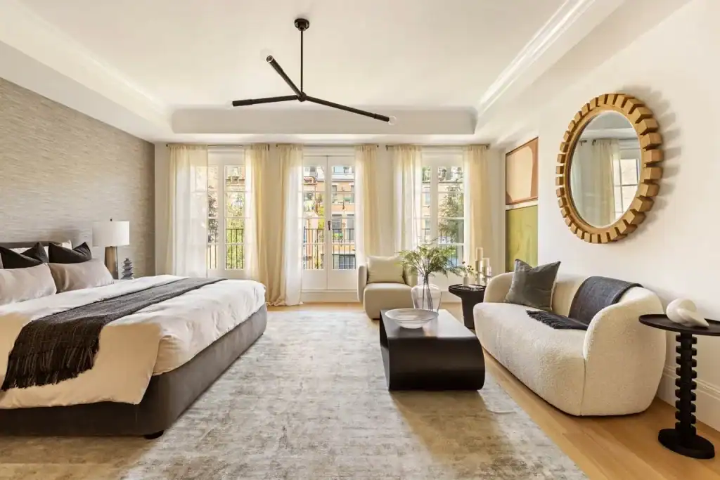 An impeccably designed primary suite in a landmark systems NYC residence, bathed in soft daylight filtering through three sets of French doors with sheer curtains, featuring a low-profile upholstered bed with charcoal accents, a textured accent wall, a sculptural black chandelier, a curved ivory bouclé sofa, a gold-framed mirror, and a minimalist coffee table creating a refined modern atmosphere