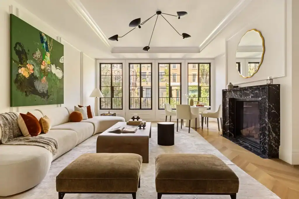 A sprawling open-concept living space in a landmark systems NYC residence, illuminated by tall black-framed French windows with views of a leafy urban backdrop, blending modern comfort with classic architectural details, including a curved cream sectional with burnt-orange accents, a forest-green abstract painting, a black marble fireplace with a gold-framed mirror, light-oak chevron floors, and a sculptural chandelier under a tray ceiling