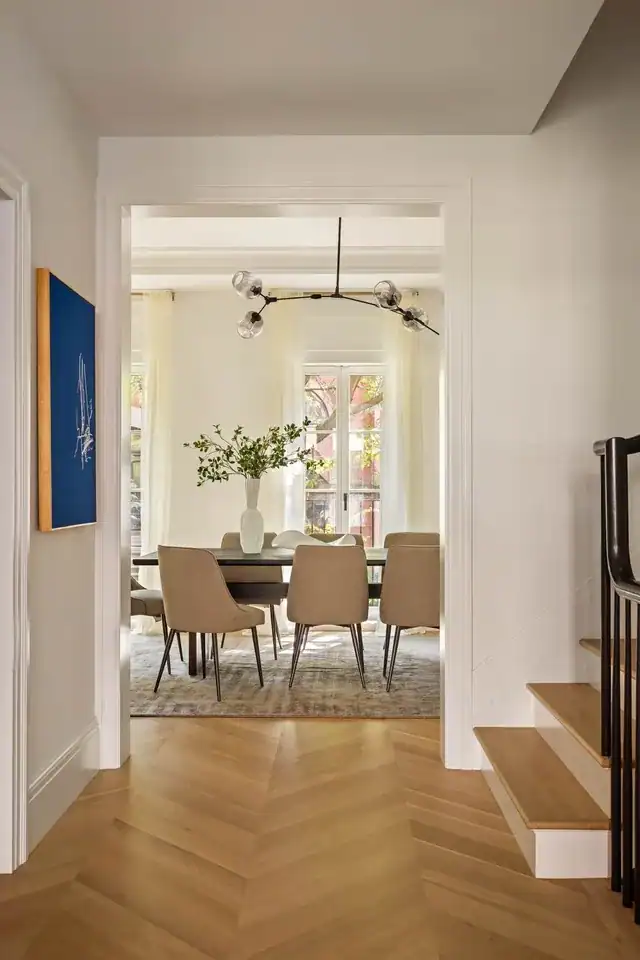 An elegant, light-filled dining room, glimpsed through a wide white doorway from a hallway with light-oak chevron-patterned floors, features a dark wood table surrounded by tan upholstered chairs on a textured gray area rug, a modern branch-like chandelier with smoked glass globes suspended from the ceiling, and tall windows draped in sheer white curtains that frame views of a brick exterior and lush greenery, while a vibrant cobalt blue abstract painting on the left wall and a minimalist staircase on the right add contemporary flair to the sophisticated space.