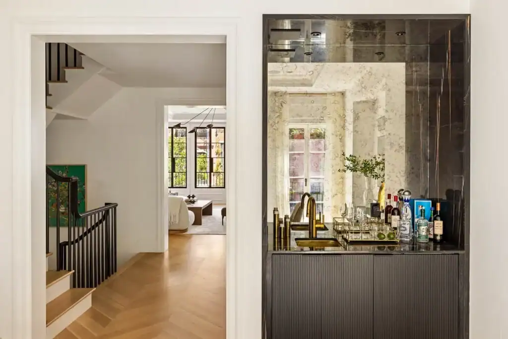 Landmark Systems NYC luxury home bar, black marble countertop, integrated gold sink and faucet, white recessed wall, antiqued mirror, reflected window light, open doorway, light-oak chevron hallway, black wrought-iron staircase railing, sun-drenched living room, high-end interior design.