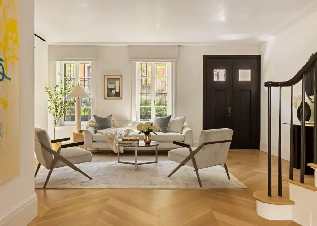 An exquisitely appointed, sun-drenched living space, characterized by its luminous atmosphere and sophisticated neutral palette, features a masterfully arranged conversation area anchored by a plush off-white sofa draped with a luxurious long-pile throw and two striking mid-century modern lounge chairs with geometric timber frames, all resting atop a subtle, tonal area rug on elegant light-oak chevron-patterned floors, while the composition is further elevated by a minimalist circular coffee table, a sculptural floor lamp, two expansive windows framed by understated Roman shades, a bold charcoal-hued double entrance door, and a glimpse of a refined staircase with a polished black handrail, collectively embodying a pinnacle of modern urban refinement.