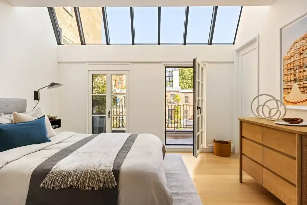 Top-floor bedroom with angled skylight, natural daylight, French doors, private balcony, urban brick views, charcoal-gray bed runner, teal accent pillow, mid-century oak dresser, architectural photo of landmark systems NYC ornate building, hardwood floors, gray area rug, minimalist modern design.