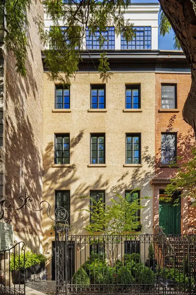 A stately multi-story townhouse, featuring a warm tan stucco facade and a series of dark-framed, multi-paned wood windows arranged in a precise grid, stands elegantly nestled between a classic red-brick neighbor and a sun-dappled tree, while a modern glass-enclosed penthouse level with blue-tinted windows sits atop the structure, all secured behind an ornate black wrought-iron fence and gate that enclose a lushly landscaped front garden filled with vibrant green shrubbery and delicate trees.