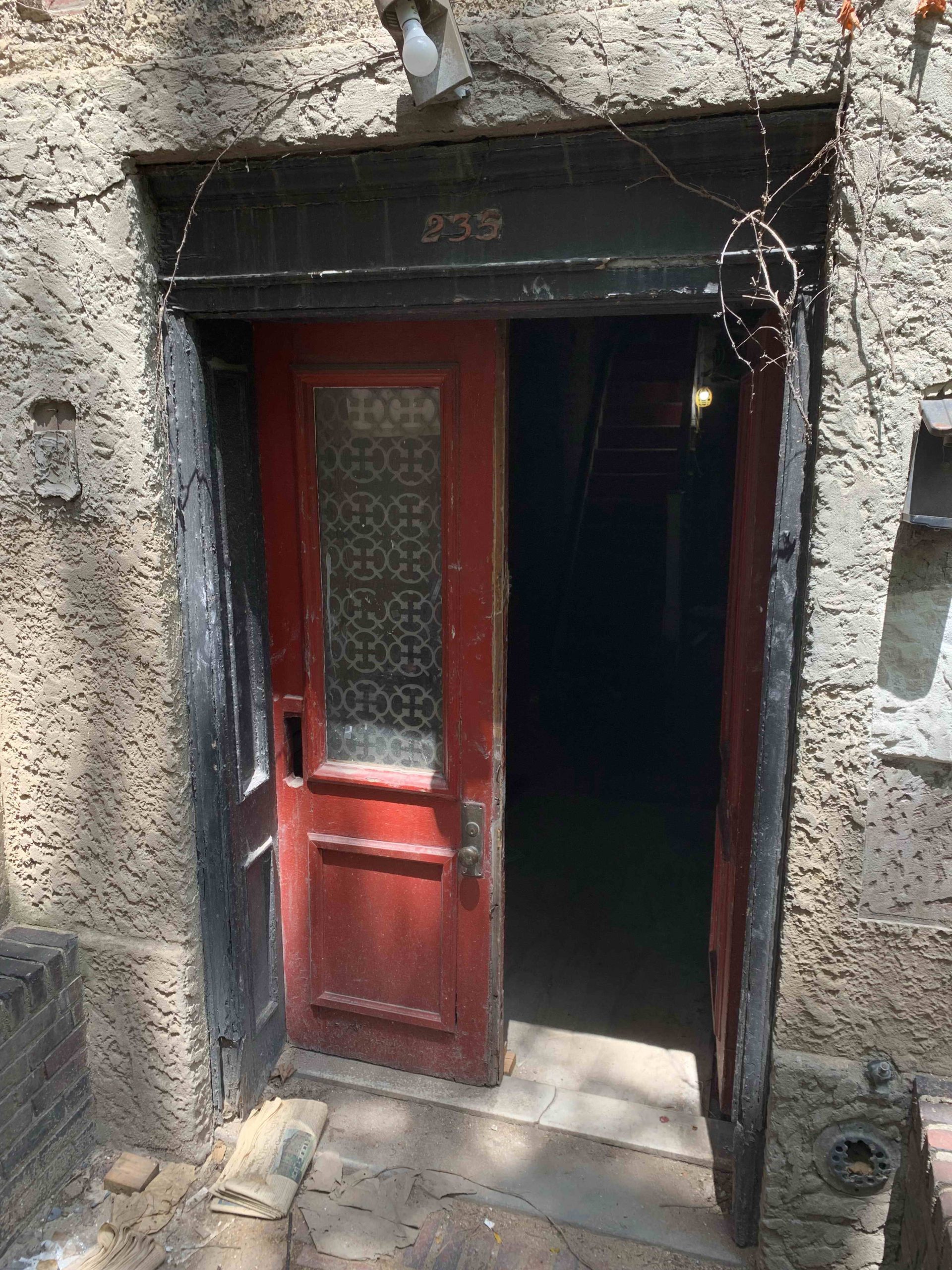 Open red wooden entrance door of an old building before renovation, showing number 233 on a dark frame and a worn stucco wall.