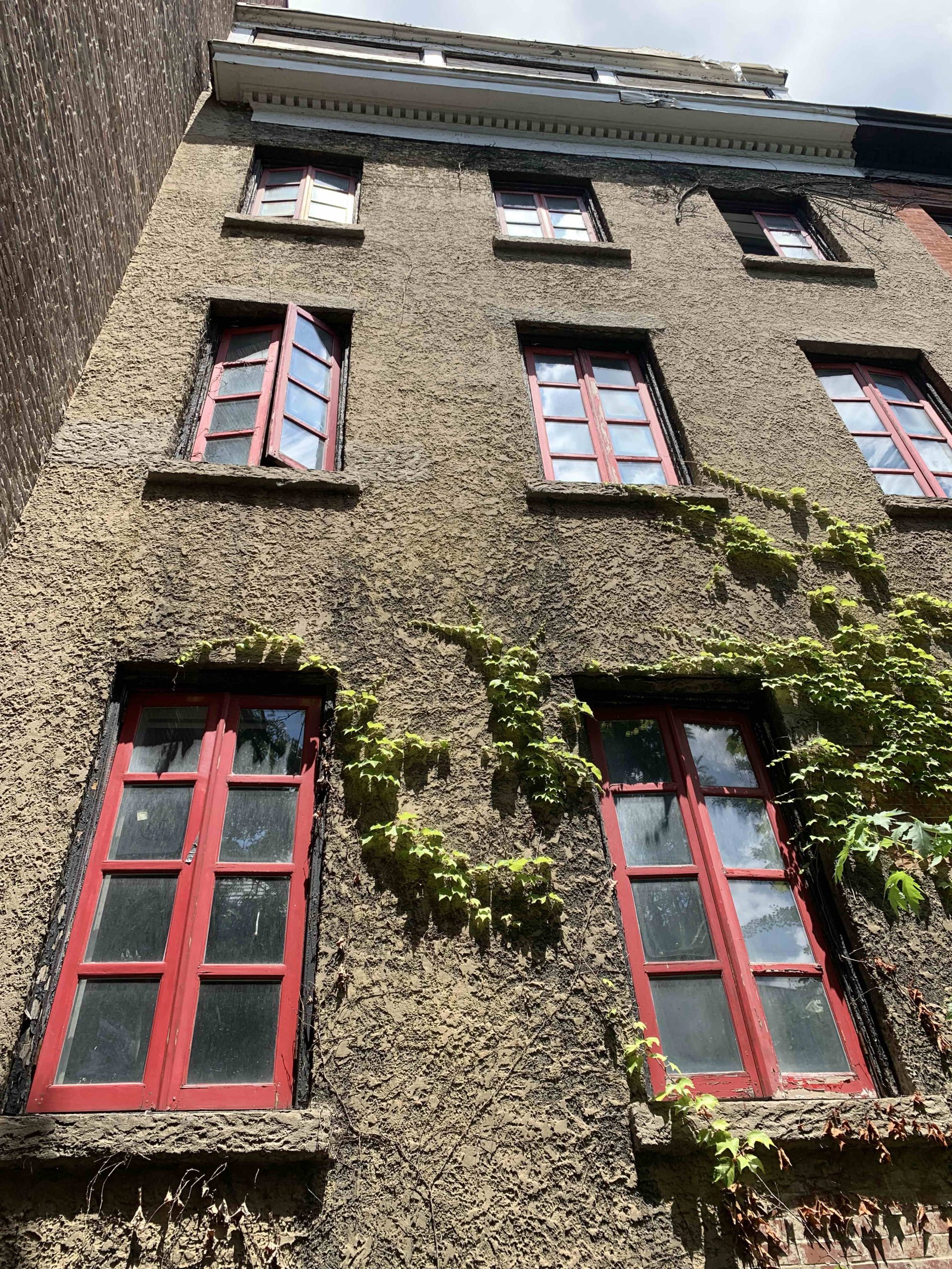 Low-angle view of an old building facade before renovation, featuring worn textured stucco walls, weathered red wooden window frames, and overgrown ivy.